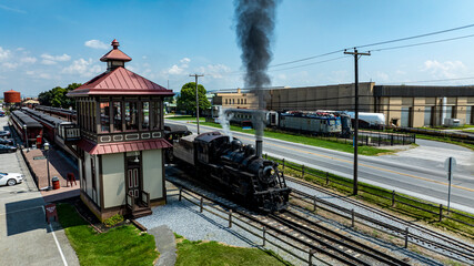 A classic steam train releases smoke as it pulls away from an old-style station under a bright blue sky. The setting captures the charm of retro transportation in an urban environment.