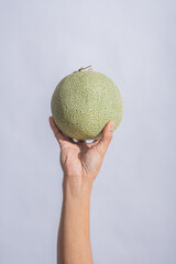 A hand holding a whole green-skinned cantaloupe melon against a bright white background, showcasing its rough textured surface, round shape, and tropical freshness of this sweet fruit