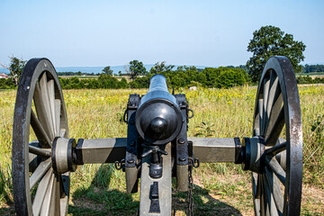 A vintage cannon stands still on a grassy knoll, pointing towards a sprawling battlefield. The sky shines blue, and trees frame the horizon, evoking a sense of history.