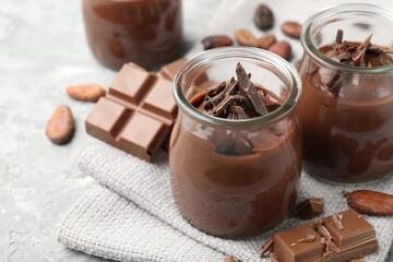 Tasty pudding in glass jars, pieces of chocolate and cocoa beans on light grey table, closeup