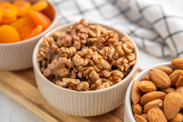 Three white ceramic bowls filled with almonds, walnuts, and dried apricots, arranged on a wooden cutting board, offer a healthy and delicious snack or ingredient for various culinary creations