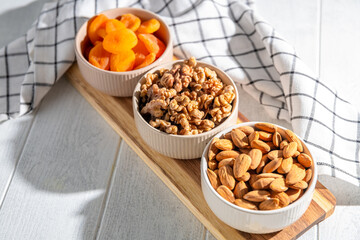 Three bowls of healthy snacks including dried apricots, walnuts, and almonds are arranged on a wooden board, offering a nutritious and delicious treat