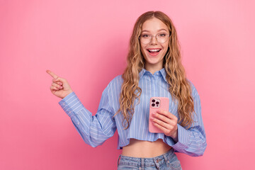 Smiling young woman holding phone and pointing joyfully against pink background wearing blue...