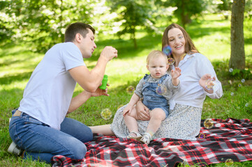 Family sitting in park during picnic, dad blowing soap bubbles, having fun playing outside, mom holding toddler in arm, laughing, catching bubbles. Playing with soap bubbles, family outdoor recreation