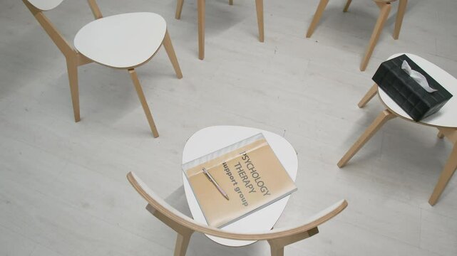 High angle tilt-down no people shot of interior of white chairs, psychology book and tissue box in minimalist group therapy room