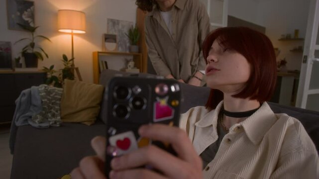 Teenage girl with smartphone sitting on couch and scrolling through shorts while her mother passing by and starting to scold and take away mobile phone of her daughter