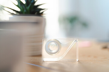 A clear tape dispenser sits on a desk in a bright office. A potted plant is blurred in the background. The image evokes productivity and minimalism.