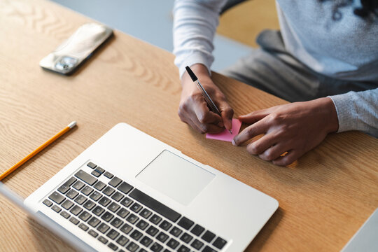 A young Arab man writes on a pink sticky note at his desk. He's working on his laptop, with a phone and pencil nearby.  He's focused on his task.