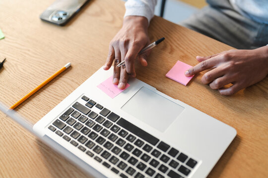 A young Arab man works at his desk, using sticky notes on his laptop. He holds a pen in one hand and points at a note with the other. A phone, pencil, and extra note are also on the desk.
