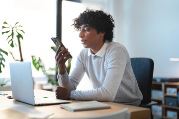 A young Arab man uses his phone's speakerphone function while working at his desk. He sits with his laptop open in a bright, modern office. He is focused on his conversation.