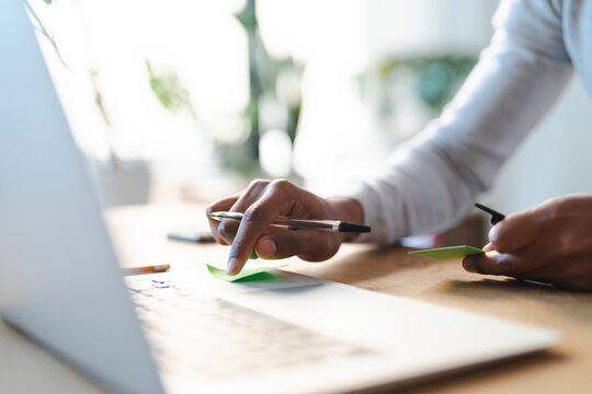 A young Arab man uses sticky notes while working on his laptop. He holds a pen in one hand and a sticky note in the other.  He is focused on his work.