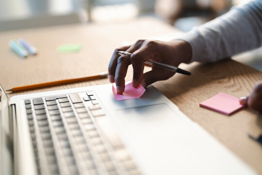 A young Arab man places a pink sticky note on his laptop. He is working at a desk with a pen, pencil, and other sticky notes nearby.  He is focused on his task.