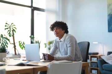 Young Arab man sits at his desk with a laptop. He is looking thoughtfully out the window in a bright, modern office space.