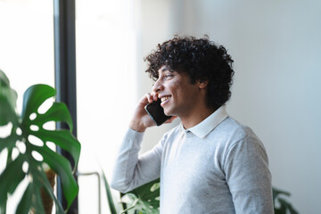 An Arab young man is smiling while talking on his smartphone. He is standing near a window with a plant.