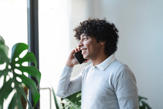 An Arab young man is smiling while talking on his smartphone. He is standing near a window with a plant.