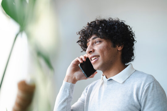 An Arab young man is smiling while talking on his smartphone. He is standing near a window with a plant.