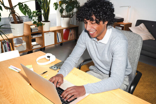 An Arab young man is working on his laptop at home. He is smiling and seems to be enjoying his work in a bright, plant-filled space.