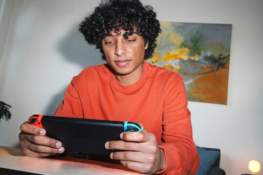 An Arab young man is focused while playing a handheld video game console. He is wearing an orange sweater and sitting at a table.