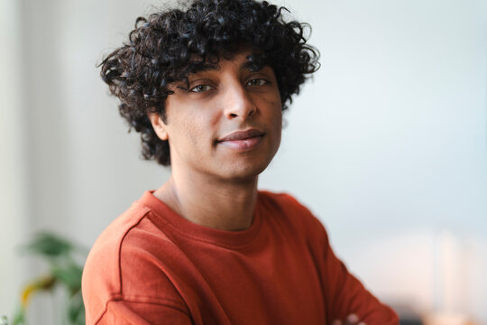 Portrait of a young Arab man with curly hair. He is wearing a casual orange shirt and looking directly at the camera.