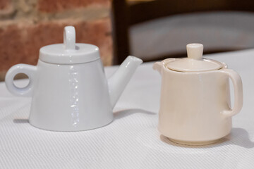 Two teapots, one white and one beige, placed on table with rustic brick wall in background, creating a cozy and inviting atmosphere