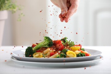 Woman sprinkling spices onto pasta with vegetables at white table indoors, closeup