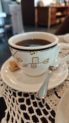 Cup of black coffee served on a saucer with a spoon and small biscuits on a crocheted tablecloth. Concept of morning routine, cozy cafe moments and warm beverages