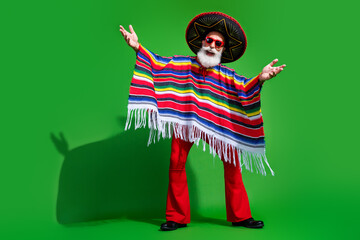 Festive mature man in traditional Mexican outfit with colorful poncho and sombrero posing against a green background