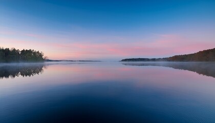 Fototapeta premium A calm lake reflects pink and blue sky hues at dawn or dusk. Tree lined shores feature dense greenery on one side and autumn tones on the other, with light mist over the water.