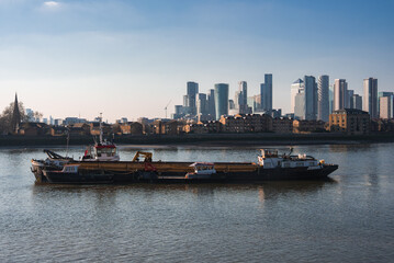 Fototapeta premium A barge floats on the River Thames with Canary Wharf's modern skyscrapers and a church spire visible, reflecting on calm water under a clear blue sky.