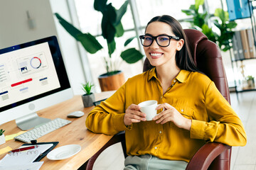 Smiling professional businesswoman enjoying coffee in a modern office workspace with daylight and...
