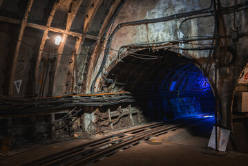 Aged underground tunnel in London with exposed concrete walls, structural reinforcements, and tracks leading into a dimly lit section with blue light.