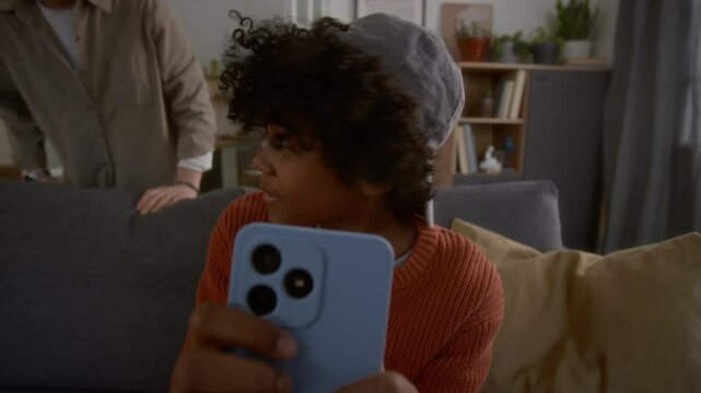 Cute African American boy sitting on couch and playing video game or texting in smartphone while his nother coming up to him and starting to scold and take his phone away