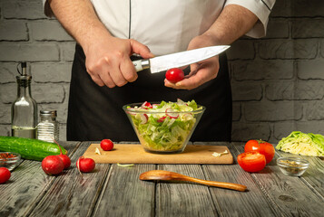 Chef cuts radishes while preparing fresh salad in modern kitchen. Various vegetables and seasonings are laid out on rustic wooden table.