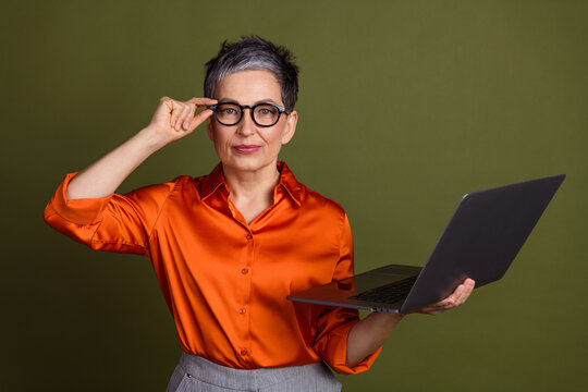 Confident mature professional holding a laptop and adjusting eyeglasses against a green background in a vibrant orange shirt