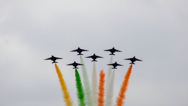 Aerial display of fighter jets leaving vibrant orange, white, and green smoke trails in the sky, representing the Indian flag. The planes fly in a precise V-formation against a pale sky.

 - Powered by Adobe