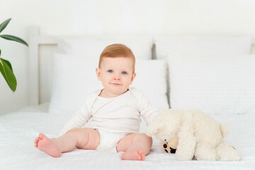 Fototapeta premium A cute little smiling baby boy in a white bodysuit is sitting on a bed at home in a bright bedroom with a soft teddy bear, a six-month-old baby with blue eyes is smiling or laughing