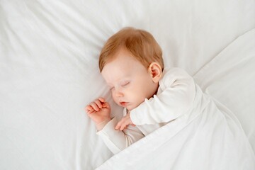 Baby boy sleeping under a blanket in a white bodysuit on the bed with his hand folded under his...