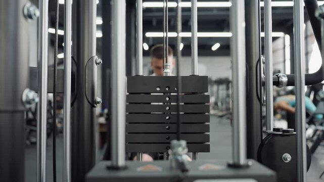 View through vertical steel rods of cable machine as athlete seated on bench pulls weight stack handle in controlled rowing movement showcasing strength and focus in bright modern gym environment