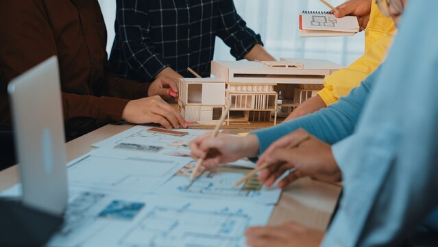 A diverse team engages in brainstorming and planning an architectural project. The scene showcases hands working on models and drawings in a creative workspace environment. SACTR