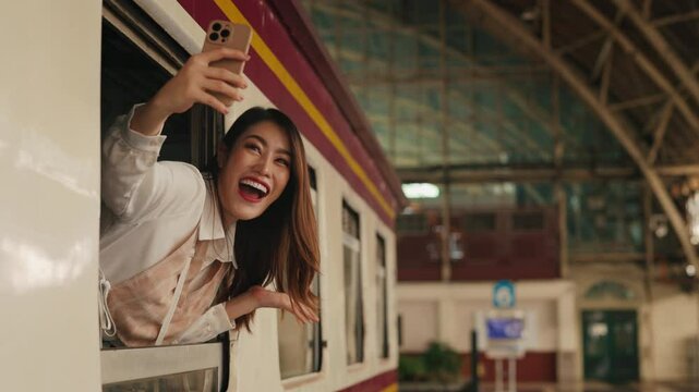 Portrait of joyful Asian trans woman taking selfie on smartphone leaning out of railroad car window. Happy female takes picture for social media at train station. Funny travel