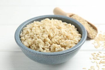 Tasty brown rice in bowl and raw grains on white wooden table, closeup