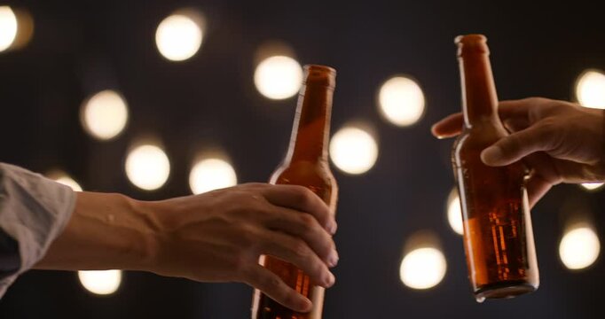 Beer splashing from clinking bottles in male hands during cheerful toast warm lights in background closeup celebration of fun friendship spontaneity and night party lifestyle moment with motion