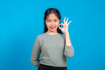 Fototapeta premium Confident young Asian woman showing an OK hand gesture while smiling slightly and winking. Her playful and approving facial expression stands out against the solid blue background.