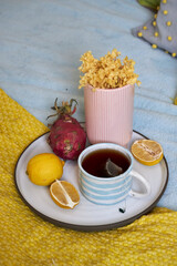 A tray with tea, flowers, and fruit placed on a bed with bright yellow, pink, blue, and gray textiles — breakfast in bed concept.