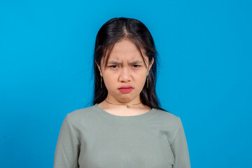 Obraz premium Close-up portrait of an angry young Asian woman frowning intensely, lips pressed and eyebrows furrowed, expressing frustration or irritation. Studio shot on solid blue background.