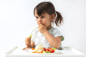 A little girl sitting in a high chair and eating fresh vegetables. She is focused while tasting food, holding a piece of celery in her hand. Concept of healthy eating and childhood nutrition