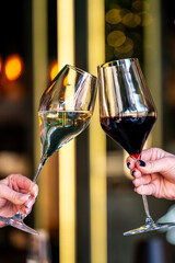 Two hands clinking elegant wine glasses, one filled with white wine and the other with red. A warm, festive atmosphere is created with blurred lights in the background.
