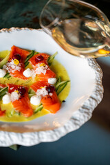 A gourmet dish featuring sliced smoked salmon arranged elegantly with cucumber and garnished with dollops of cream. A glass of white wine is blurred in the background.