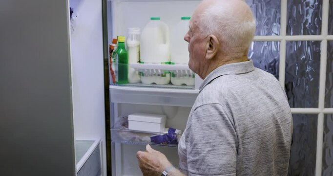 Fridge, food and home of retirement man eating grapes for snack, health and wellness nutrition. Fruit, hungry and elderly man with appetite searching and thinking of meal idea in kitchen at night.
