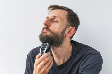 Young man aged 30 35 trimming beard with electric trimmer at home, focused expression, casual clothing, white background, close-up view of personal grooming routine concept of lifestyle